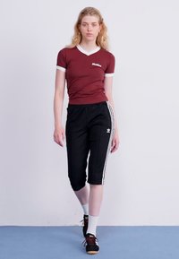 Top corto bordeaux a maniche corte con bordi bianchi, abbinato a pantaloni neri a metà polpaccio con due strisce laterali bianche e un piccolo logo. Sneakers nere e calzini bianchi.