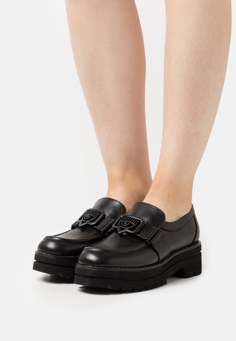 Chaussures mocassins noires épaisses avec semelles épaisses et boucles décoratives portées pieds nus sur fond blanc.