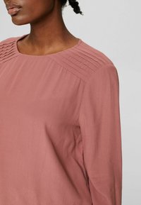 Langärmlige Bluse aus weichem Rosastoff mit gefälteltem Detail an den Schultern und Rundhalsausschnitt. Glatte Textur und lockere Passform.