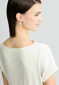 Bijela, kratka majica s kratkim rukavima, širokim izrezom i mekanom teksturom. Ima suptilan detalj šava duž leđa. Glatki, minimalistički dizajn.