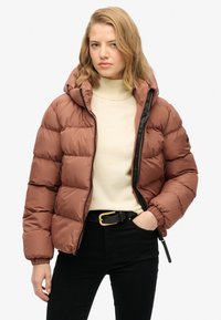 Chaqueta de plumón marrón con capucha, con un acabado brillante, cierre de cremallera y puños elásticos. Llevada sobre un suéter de cuello alto crema, combinada con pantalones negros.