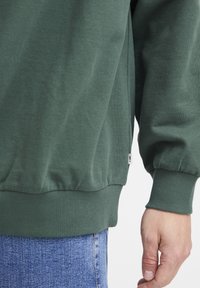 Sudadera verde hecha de suave algodón con puños y dobladillo acanalados; presenta un acento en la costura lateral y una pequeña etiqueta de marca.