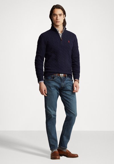 Marineblå kabelstrikket sweater med halv-lynlås krave, parret med blå denim jeans og brune lædersko. Har kontraststribede skjorteærmer.