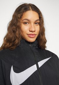 Mustat Nike-takki, jossa on korkea kaulus, vetoketjusuljenta ja suuri valkoinen swoosh-logo. Rakenteellinen kangas, jolla on kevyt ulkonäkö.