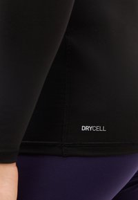 Μαύρο αθλητικό τοπ με εμφανείς ραφές και λογότυπο «DRYCELL», φορεμένο πάνω από σκούρο μωβ κολάν στο πλευρό ενός ατόμου.