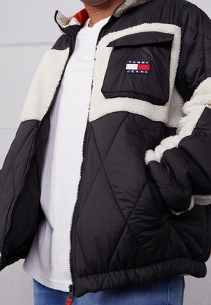 Persona che indossa una giacca trapuntata bianca e nera con dettagli in shearling e una toppa con il logo Tommy Jeans sulla tasca del petto, sopra una camicia bianca.