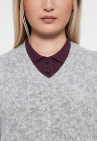 Šedý pletený sveter s výstrihom do V, so štruktúrovaným vzorom a vínovým košele pod ním, zapnutou dvoma gombíkmi.