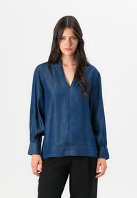 Denimskjorta i mörkblått med v-ringning och långa ärmar. Har smockade manschetter och en avslappnad passform. Slät textur med minimal sömnad synlig.