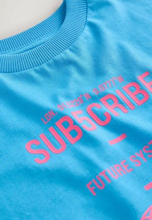 Blauer Baumwoll-Sweatshirt mit pinkem Text: "LDN. 51.5229° N 0.0777° W SUB5CRIBE FUTURE SYSTEM." Ribkragen und glatte Textur.