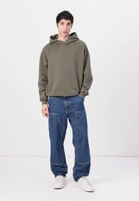 Olive green hoodie with a kangaroo pocket, paired with loose-fitting blue jeans featuring prominent seams and white sneakers.