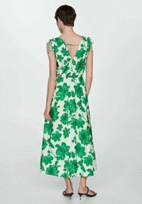 Maxi haljina u bijeloj boji s velikim zelenim cvjetnim uzorcima, s podesivim naramenicama i V-izrezom na leđima. Nosene ravne sandale.