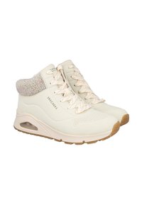 Zapatillas altas de color crema con un cuello de punto texturizado, que presentan una suela acolchada, diseño perforado y suela de goma marrón claro.