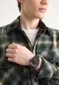 Ceas hexagonal negru cu o brățară texturată și marcaje albe, purtat pe o mână, însoțit de o brățară din mărgele negre, pe un fundal cu o cămașă în carouri.