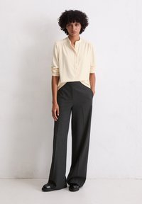 Lichtbeige, lange mouwen shirt met een bandkragen en knoopsluiting, gecombineerd met donker gestreepte, wijde broek en zwarte schoenen.