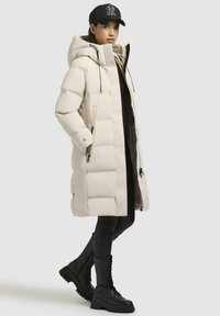 Piumino beige con colletto alto, tasche con zip e una texture lucida. Indossato sopra leggings neri e stivaletti neri massicci. Accento con berretto.