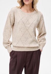 Beżowy sweter dzianinowy z wzorem diamentowym, okrągłym dekoltem i bufkami na rękawach; zestawiony z ciemnobrązowymi spodniami.