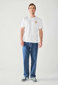 Ung mand står, iført hvid grafisk t-shirt, blå jeans og hvid-grønne sneakers foran en enkel lys baggrund.