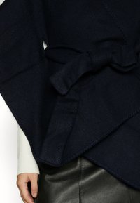 Gros plan d'un vêtement cache-cœur bleu marine noué en un nœud sur un haut blanc à manches longues et une jupe en cuir noir.