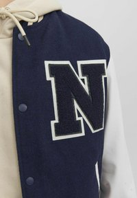 Marineblauwe en witte varsity jack met een getextureerde letter 'N' patch, drukknoopjes en een gladde witte leren mouw tegen een beige hoodie.