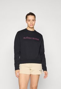 Sweatshirt preta com punhos e cós canelados, com "BJÖRN BORG" em letras cor-de-rosa a atravessar o peito. Combinado com calções bege.