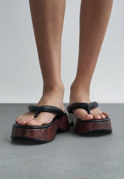 Nohy v černých kožených sandálech typu thong s tlustými, zakřivenými tmavě hnědými platformními podrážkami stojí na šedé podlaze.