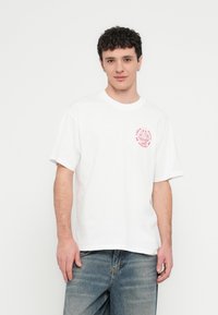 T-shirt branca de algodão com decote redondo, mangas curtas e estampa gráfica rosa na área esquerda do peito. Corte clássico, textura suave.