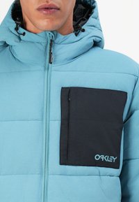 Uomo in giacca imbottita azzurro chiaro con cappuccio, cerniera frontale e grande tasca nera sul petto con logo Oakley.