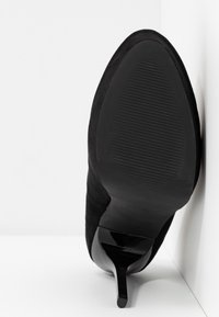 Chaussure à talon haut en daim noir avec un bout pointu et un fin talon aiguille. La semelle présente des rainures horizontales pour une meilleure adhérence.