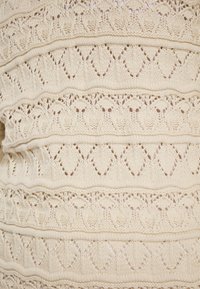 Beige gebreide stof met complexe patronen van diamanten en bloemmotieven. De textuur is zacht met open steken die een ademend ontwerp creëren.