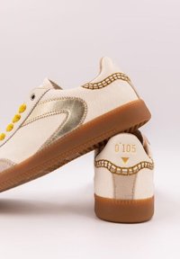 Sneakers bianchi in pelle con dettagli dorati, occhielli gialli e suola testurizzata color cammello. Presentano cuciture decorative e logo sul tallone.