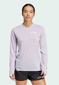 T-shirt de sport à manches longues en lavande clair avec un col ras du cou et des panneaux en maille sur les côtés, portant un logo "TERREX" blanc. Associé à un short noir.
