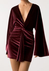 Robe mini en velours bordeaux avec un profond décolleté en V, des manches longues évasées et un détail de nœud à l'avant, présentant une texture lisse et douce.