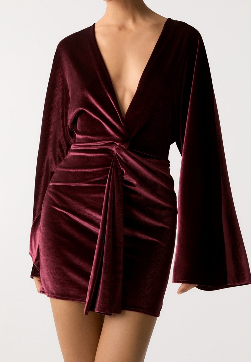 Robe mini en velours bordeaux avec un profond décolleté en V, des manches longues évasées et un détail de nœud à l'avant, présentant une texture lisse et douce.
