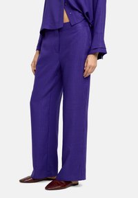 Spodnie o szerokich nogawkach w żywym odcieniu fioletu, wykonane z teksturowanej tkaniny, posiadają płaski przód i boczne kieszenie, zestawione z prostymi brązowymi butami wsuwanymi.