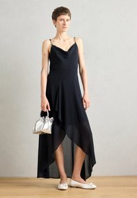 Rochie asimetrică neagră cu design drapat și accente metalice pe bretele. Modelul ține o geantă argintie și poartă adidași crem pe un podea din lemn.