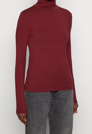 Personne portant un col roulé moulant rouge foncé à manches longues et un jean noir délavé, debout devant un fond clair uni.