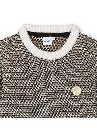 Strikket genser med et teksturert mønster i svart, krem og beige, med hvit ribbestrikket krage og en liten logoemblem foran.