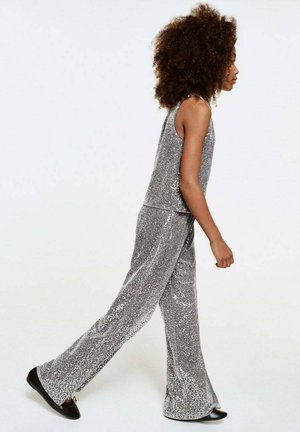 Shiny silver sequin outfit with a sleeveless top and flared pants. Model wears black shoes, showcasing a textured and glimmering design.