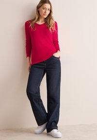 Haut rouge à manches longues avec un col rond, associé à un jean bleu foncé à larges jambes. Baskets blanches, texture lisse et design simple.