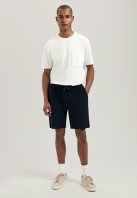 Hvit bomulls t-skjorte med rund hals og brystlomme, kombinert med mørkeblå, teksturerte, shorts med elastisk linning og beige joggesko.
