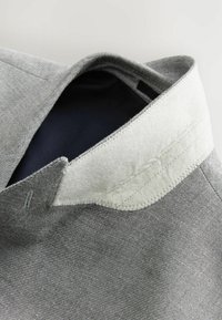 Chemise à col en tissu texturé gris avec un col en feutre blanc. Présente des coutures apparentes et une doublure intérieure sombre.