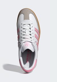 Biely športový kožený topánok s ružovými semišovými detailmi a tromi pruhmi. Má textúrovanú sivú špičku, podrážku z gumy a biele šnúrky.