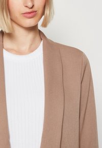 Béžový, strukturovaný blazer s otevřeným předním designem přes bílý žebrovaný top. Tkanina vypadá měkce s hladkým povrchem.