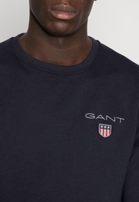 Sudadera de algodón azul marino con cuello redondo, que presenta un logo blanco de "GANT" y un emblema de escudo con acentos rojos y azules.