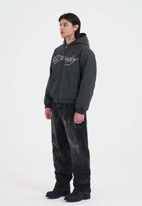 Uomo con tatuaggi sul collo e sulle mani, che indossa un cappuccio nero Ed Hardy, jeans neri stampati e stivali neri, in piedi contro uno sfondo bianco semplice.