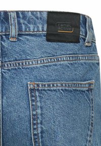 Close-up van de achterzak van een blauwe spijkerbroek met een zwart leren merklabel van Camel Active op de tailleband.