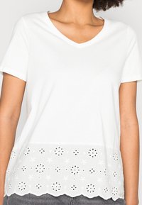 T-shirt bianco con scollo a V e motivo floreale ricamato a occhioletto, orlo ondulato e materiale in cotone, dotato di maniche corte e vestibilità rilassata.