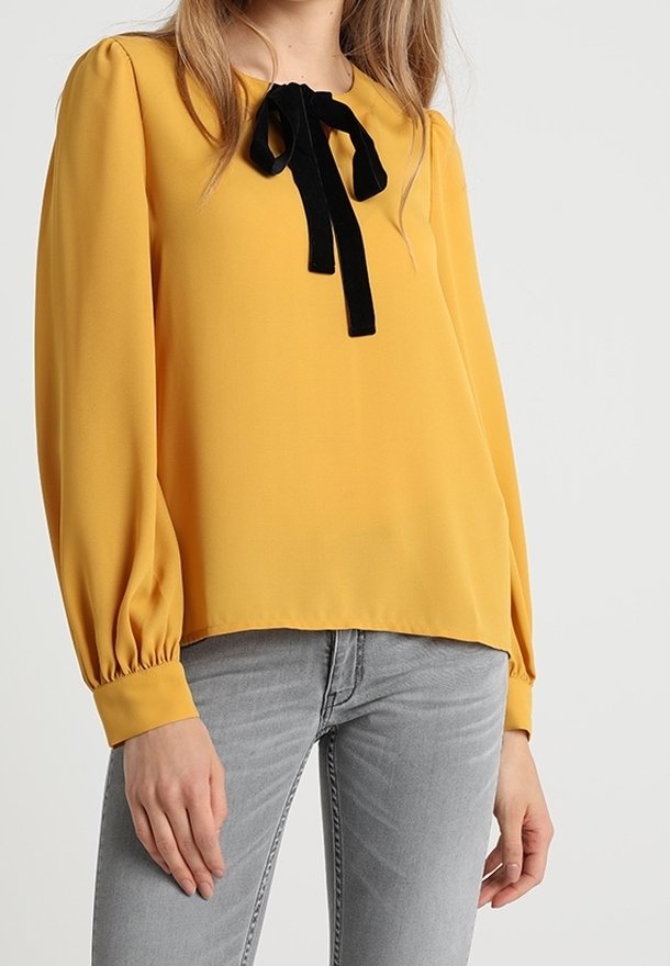 Femme portant un chemisier à manches longues jaune moutarde avec un nœud noir au col, associé à un jean gris clair.