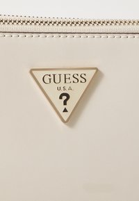 Bijela novčanik od umjetne kože s trokutastim krem simbolom koji sadrži "GUESS U.S.A." u crnom tekstu i simbolom upitnika. Detalj zatvaranja na munje.