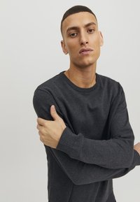 Suéter gris de punto con cuello redondo, puños acanalados y un corte holgado. El material parece suave y texturizado. Modelo con los brazos cruzados.